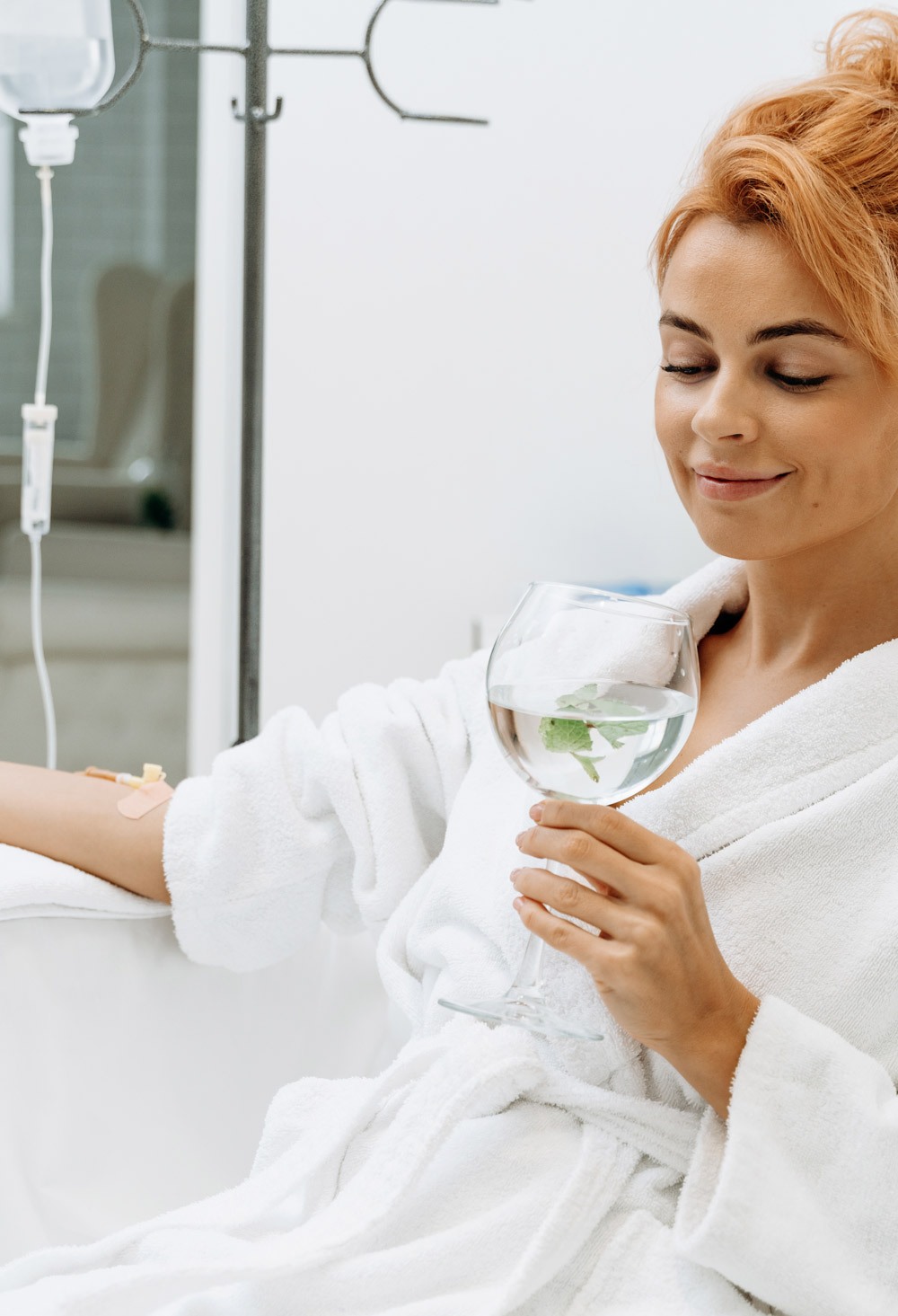 Waist up portrait view of the charming woman in white bathrobe sitting in armchair and receiving IV infusion. She is holding glass of lemon beverage and smiling Waist up portrait view of the charming woman in white bathrobe sitting in armchair and receiving IV infusion. She is holding glass of lemon beverage and smiling