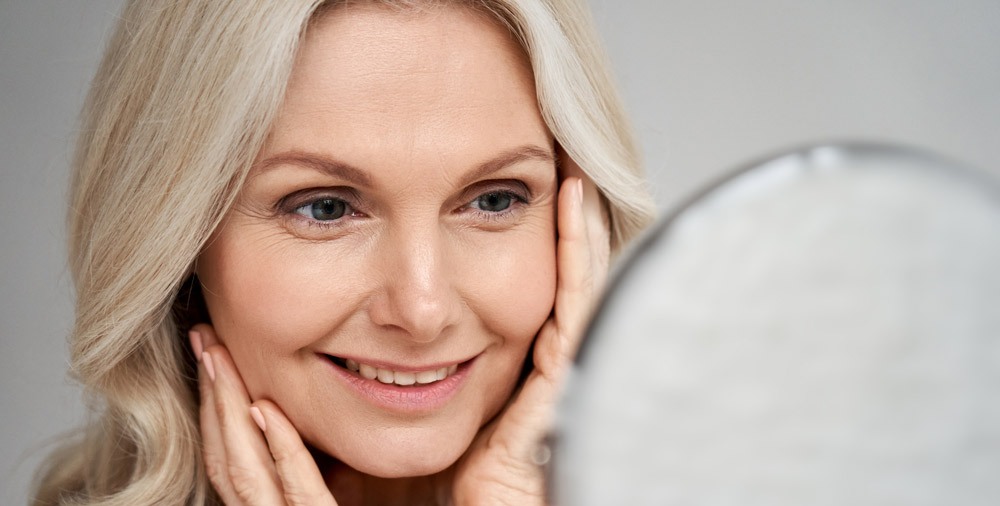 Happy 50s middle aged woman model touching face skin looking in mirror. Smiling mature older lady pampering, enjoying healthy skin care, aging beauty, skincare treatment cosmetic products